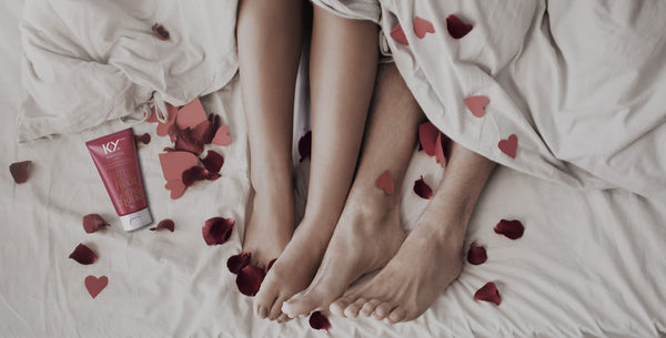 A couple’s bare feet intertwined on a white bed surrounded by scattered rose petals, with a tube of K-Y Warming Jelly placed beside them.