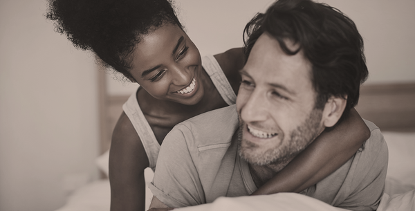 Neutral-toned photo of a woman hugging a man from behind while they relax together in a cozy bedroom.