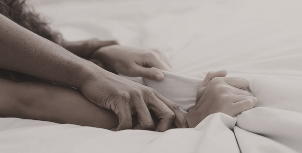  Neutral-toned close-up of a man holding a woman's wrists down on white bed sheets, demonstrating an intense intimate moment.