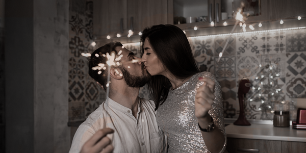 A couple kisses while holding sparklers in a cozy kitchen decorated with string lights.