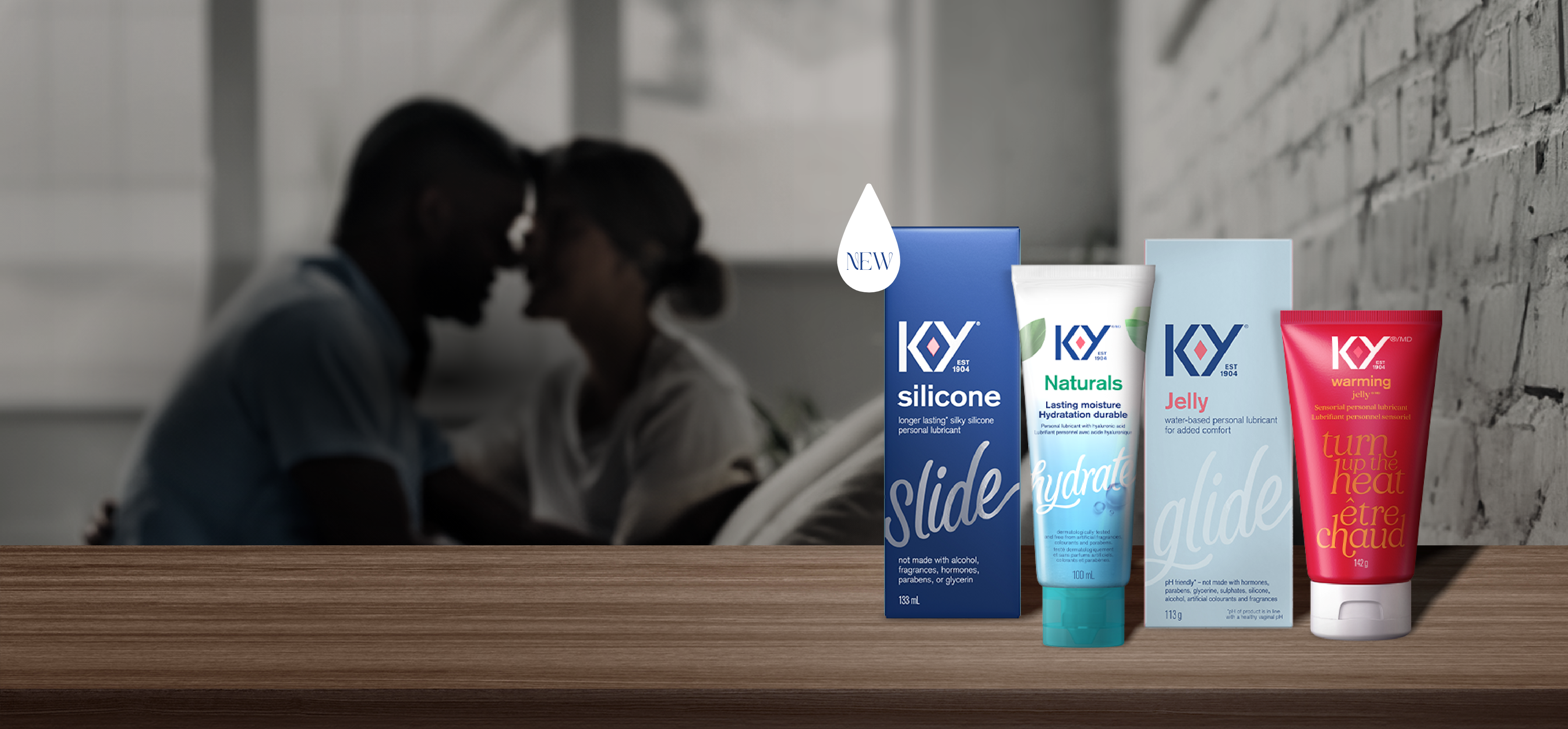 K-Y Lubricant - Silicone, K-Y Lubricant - Naturals Moisture+ Intimate Gel, K-Y Lubricant - Gel, and K-Y Lubricant - Warming Gel on a wooden shelf in front of a couple facing each other sitting on a bed.